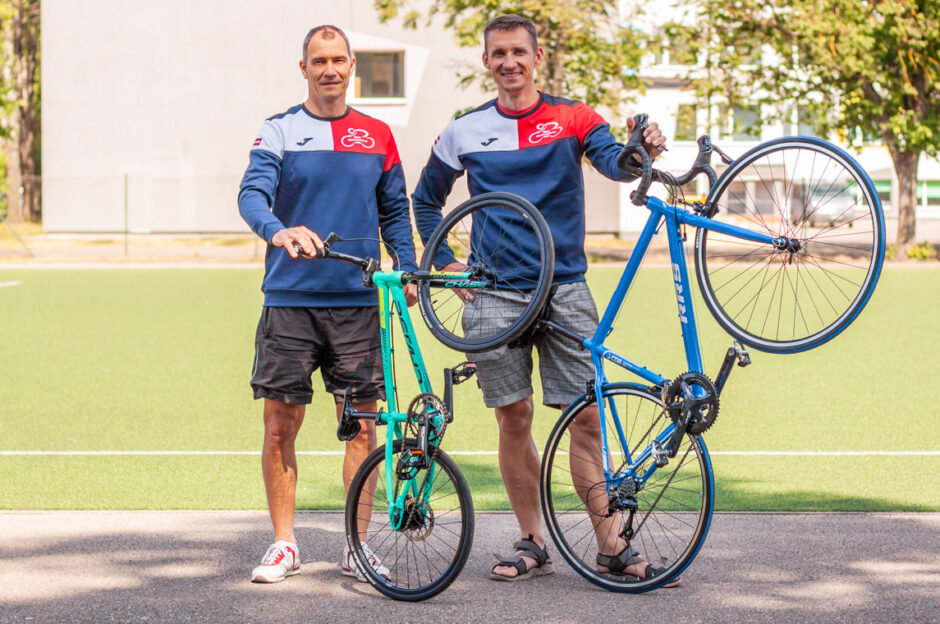 divi velobraucēji ar šosejas riteņiem pozē skolas priekšā